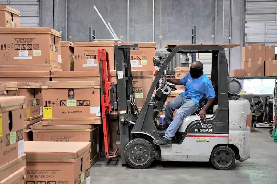 Warehouse worker operating a Nissan forklift, moving stacked cardboard boxes in a storage area while wearing a blue shirt and mask.